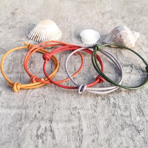 Pode incluir: Cinco pulseiras de couro coloridas em tons de laranja, vermelho, roxo e verde, cada uma com um fecho de nó. As pulseiras são exibidas com conchas em uma superfície cinza texturizada.