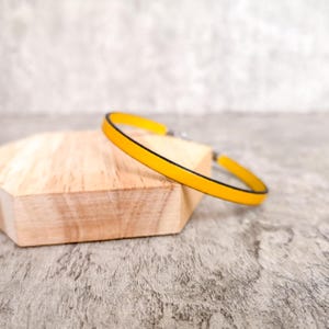 Könnte beinhalten: Ein leuchtend gelbes Armband mit schwarzem Rand liegt auf einem hellbraunen Holzblock. Das Armband hat ein schlichtes, minimalistisches Design und einen silbernen Verschluss. Der Hintergrund ist eine strukturierte graue Oberfläche.