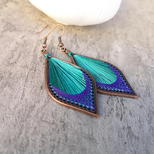 Op de afbeelding: Een paar handgemaakte oorbellen met een bladachtige vorm. De oorbellen hebben een koperkleurig frame gevuld met turquoise en paars garen. Het garen is ingewikkeld geweven, waardoor een textuurontwerp ontstaat. De oorbellen hebben een haaksluiting.