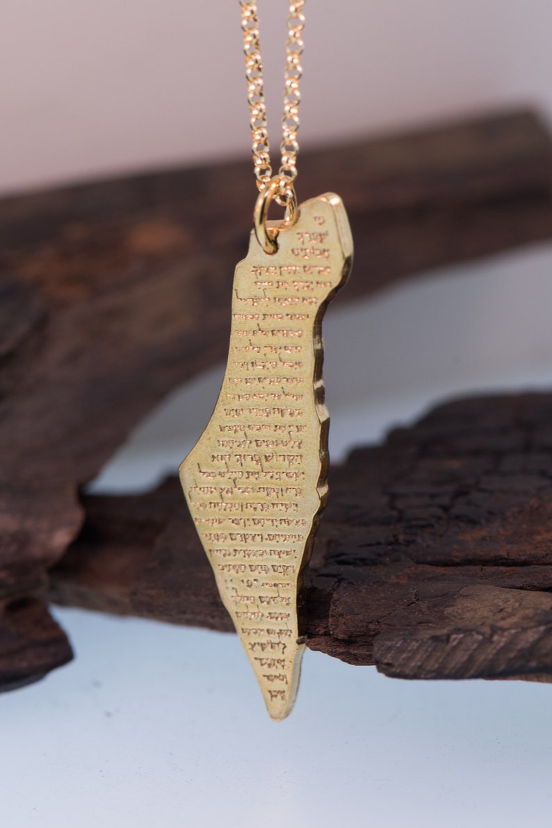 May include: A gold-colored pendant necklace featuring a map of Israel with Hebrew text. The pendant is suspended from a delicate gold chain. The necklace is displayed against a dark, textured background.