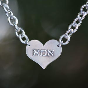 K&ouml;nnte beinhalten: Silberfarbene Halskette mit herzf&ouml;rmigem Anh&auml;nger. Der Anh&auml;nger tr&auml;gt die hebr&auml;ische Inschrift "אמא" und ist mit einer Kettengliederkette verbunden. Der Hintergrund ist ein verschwommenes, nat&uuml;rliches Gr&uuml;n.