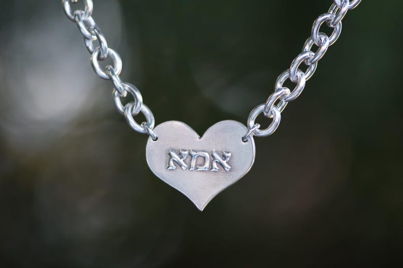 K&ouml;nnte beinhalten: Silberfarbene Halskette mit herzf&ouml;rmigem Anh&auml;nger. Der Anh&auml;nger tr&auml;gt die hebr&auml;ische Inschrift "אמא" und ist mit einer Kettengliederkette verbunden. Der Hintergrund ist ein verschwommenes, nat&uuml;rliches Gr&uuml;n.