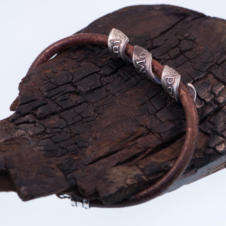 May include: A brown leather bracelet with three silver-coloured spiral beads. The beads are inscribed with the word "Mode" and other text. The bracelet is displayed on a dark, textured wooden surface.