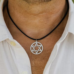 Pu&ograve; includere: Una collana con pendente color argento con un design geometrico. Il pendente &egrave; di forma circolare con un motivo intricato. La collana ha un cordino nero. La persona indossa una camicia bianca abbottonata.