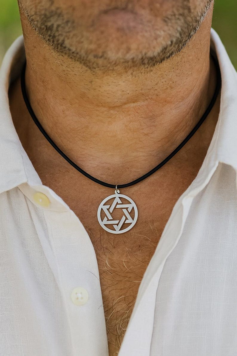Pu&ograve; includere: Una collana con pendente color argento con un design geometrico. Il pendente &egrave; di forma circolare con un motivo intricato. La collana ha un cordino nero. La persona indossa una camicia bianca abbottonata.