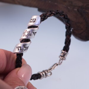 May include: A black leather bracelet with a silver charm featuring a Star of David and Hebrew text. The charm is engraved with the words "Chai" and "Shema Yisrael".