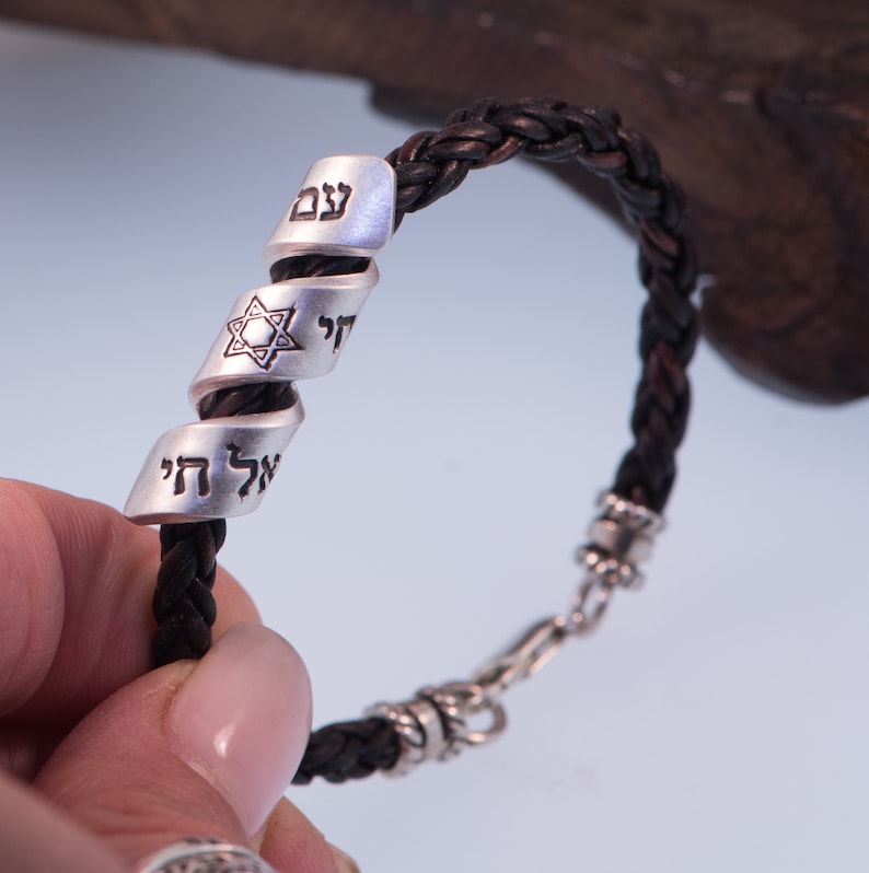 May include: A black leather bracelet with a silver charm featuring a Star of David and Hebrew text. The charm is engraved with the words "Chai" and "Shema Yisrael".