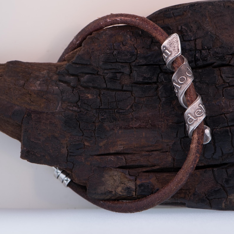 May include: A brown leather bracelet with a silver spiral design. The silver spiral has the word "model" engraved on it. The bracelet is set against a dark, textured wooden background.