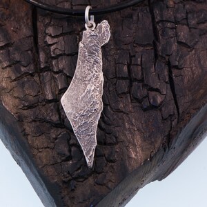 May include: A silver pendant shaped like the map of Israel with a textured, rugged surface finish. The pendant is attached to a simple silver jump ring and hangs from a thin, smooth black cord necklace, creating a contrast between the metal and cord.
