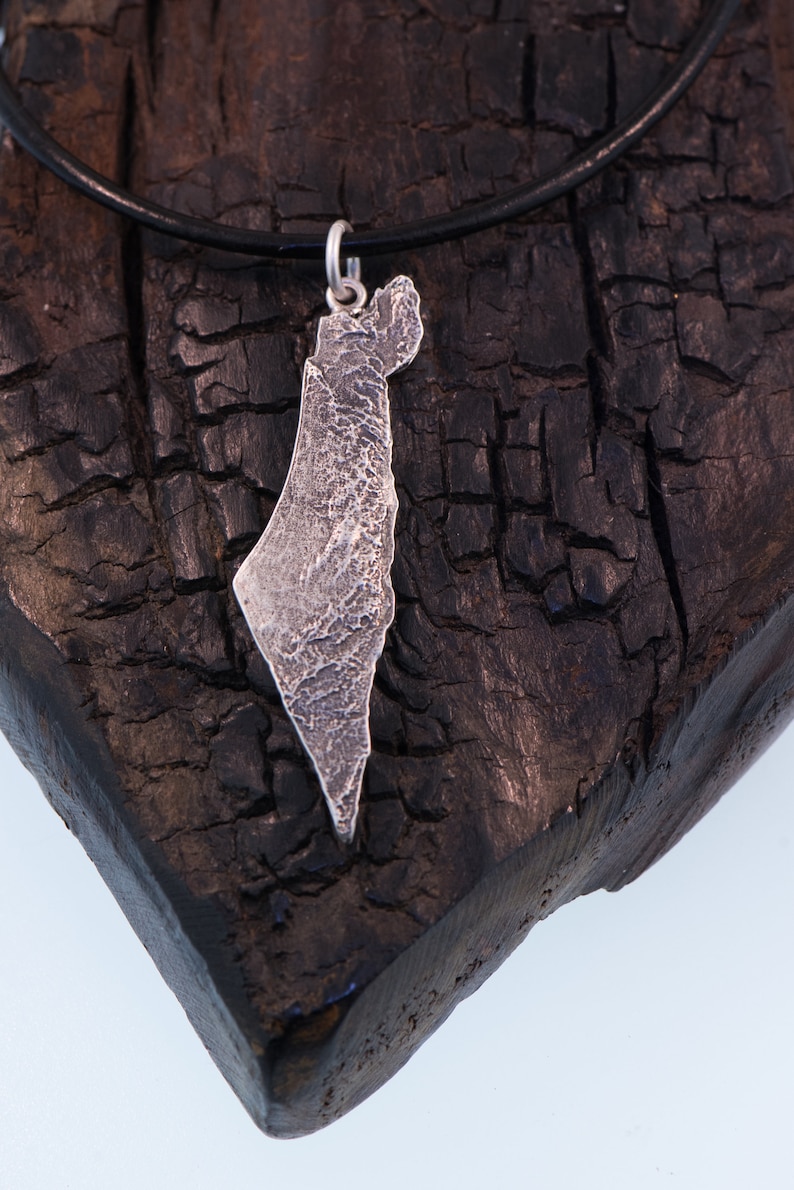 May include: A silver pendant shaped like the map of Israel with a textured, rugged surface finish. The pendant is attached to a simple silver jump ring and hangs from a thin, smooth black cord necklace, creating a contrast between the metal and cord.