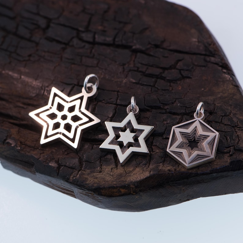 May include: Three silver star-shaped pendants with intricate designs. The pendants are displayed on a dark, textured wooden surface. Each pendant has a different star design, showcasing detailed craftsmanship and a polished finish.