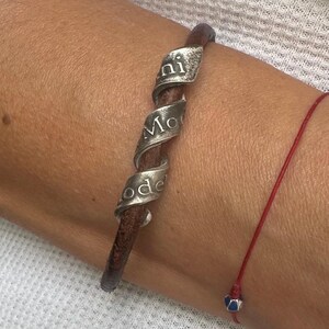 May include: A brown leather bracelet with a silver spiral design. The silver spiral has the words "ni Mo ode" engraved on it. A red string bracelet with a blue and silver bead is also visible.