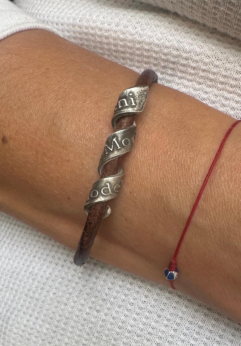 May include: A brown leather bracelet with a silver spiral design. The silver spiral has the words "ni Mo ode" engraved on it. A red string bracelet with a blue and silver bead is also visible.