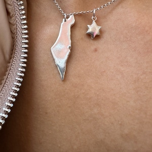 May include: Silver necklace with two pendants: a map of Israel and a six-pointed star. The pendants hang from a fine chain. The necklace is shown against a skin tone background.
