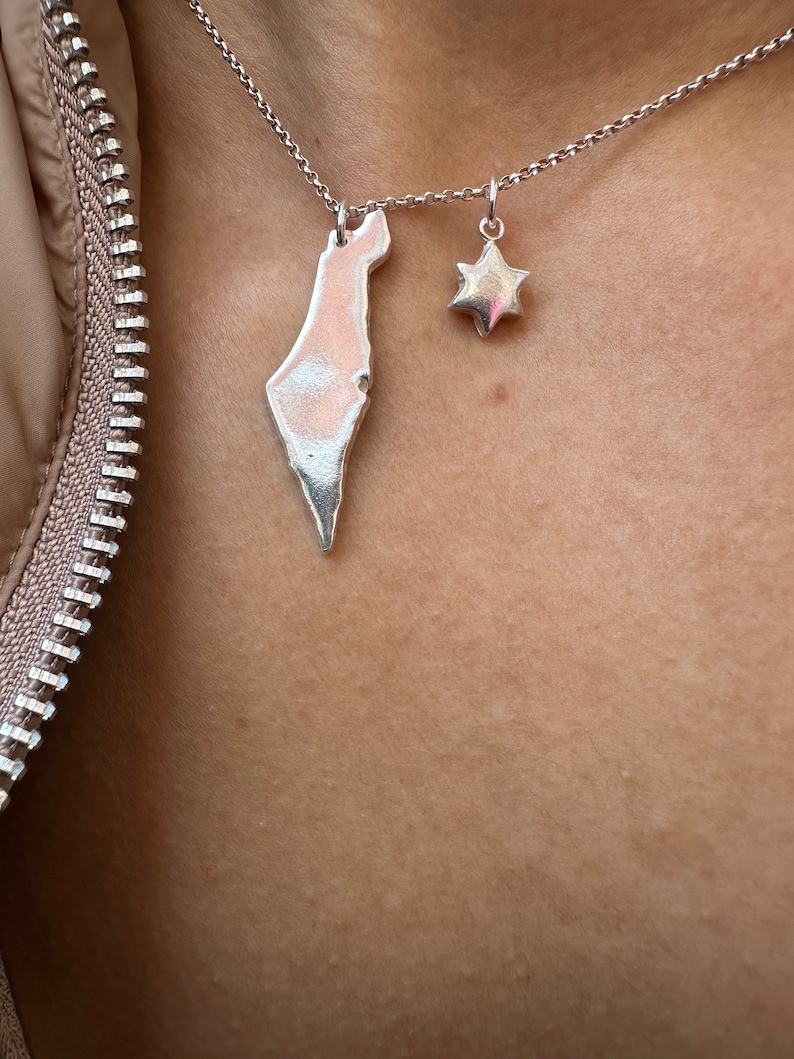 May include: Silver necklace with two pendants: a map of Israel and a six-pointed star. The pendants hang from a fine chain. The necklace is shown against a skin tone background.