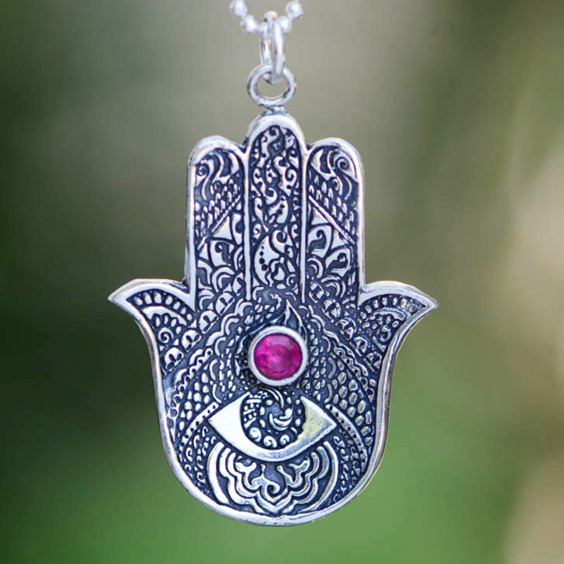 May include: Silver pendant necklace featuring a detailed hamsa hand charm with a pink gemstone eye. The hamsa hand is a symbol of protection and good luck.