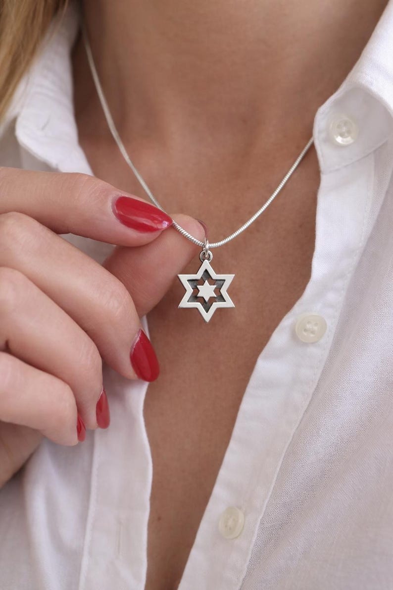 May include: Silver Star of David pendant necklace. The pendant has a six-pointed star design with a smaller star in the centre. The necklace is worn on a silver chain. The person is wearing a white button-down shirt.