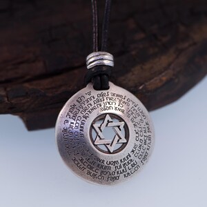 Puede incluir: Collar con colgante plateado con un dise&ntilde;o de Estrella de David en el centro. El colgante circular tiene texto grabado alrededor de la estrella. El collar tiene un cord&oacute;n negro.