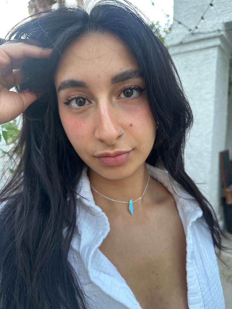 Op de afbeelding: Een vrouw met lang zwart haar draagt een wit overhemd met knopen, een zilveren kettingketting en een hanger van blauwe en groene opaal.
