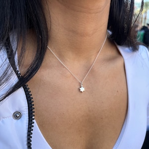 May include: A delicate silver necklace featuring a small, six-pointed star pendant. The necklace is worn against a white top. The star pendant is the central element, offering a simple and elegant design, suitable for everyday wear.