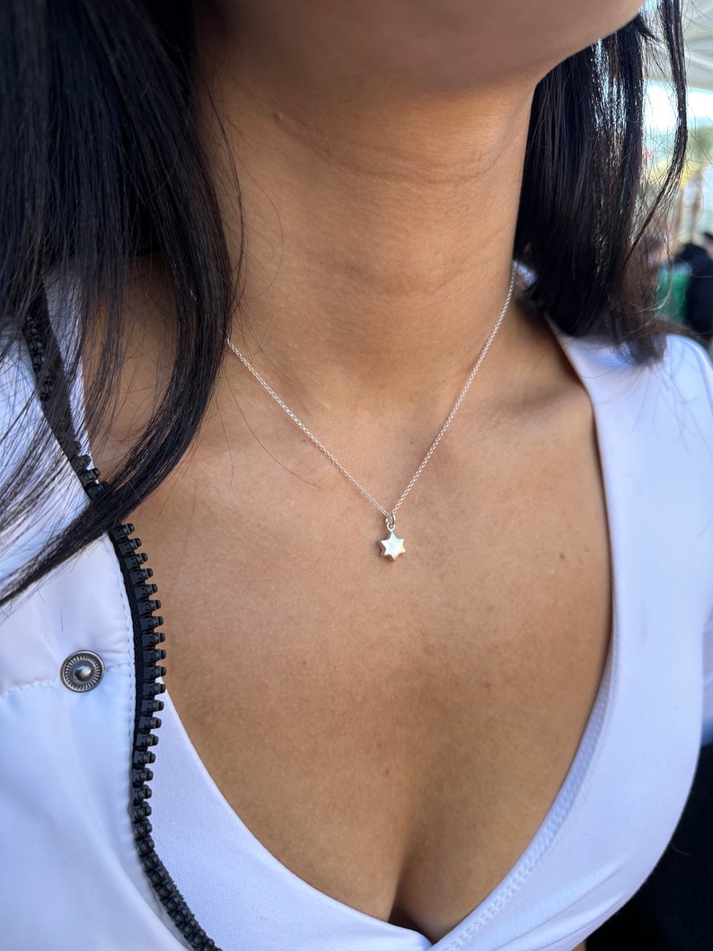 May include: A delicate silver necklace featuring a small, six-pointed star pendant. The necklace is worn against a white top. The star pendant is the central element, offering a simple and elegant design, suitable for everyday wear.
