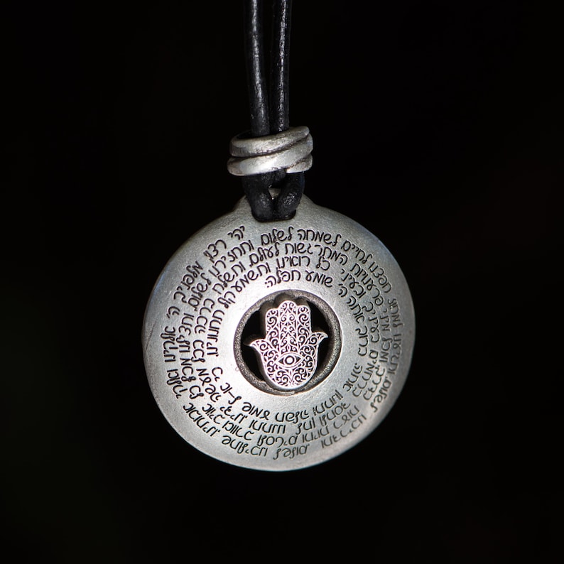 May include: A silver-colored pendant with a circular design and a black leather cord. The pendant features a central Hamsa hand symbol and is encircled by text. The background is black.