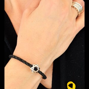 May include: A black leather bracelet with a silver Star of David charm. The bracelet is worn on a person's wrist. A yellow ribbon is pinned to the person's black shirt.