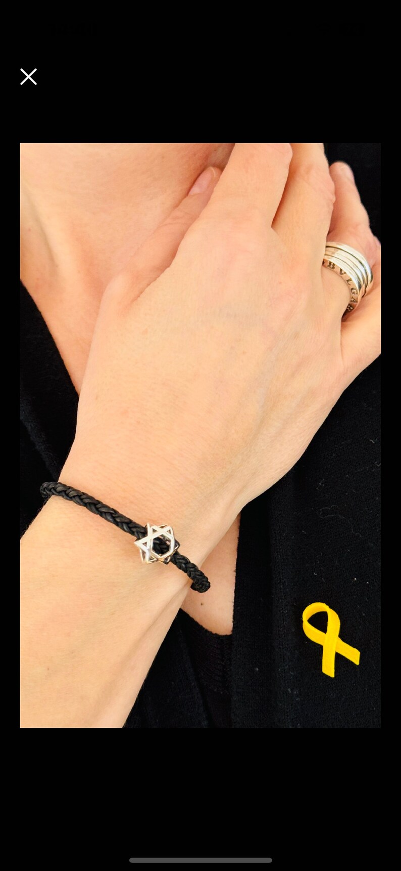 May include: A black leather bracelet with a silver Star of David charm. The bracelet is worn on a person's wrist. A yellow ribbon is pinned to the person's black shirt.