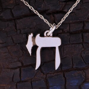 Puede incluir: Un collar de cadena de plata con un colgante de plata en forma de la letra hebrea Chai. El colgante est&aacute; sobre un fondo marr&oacute;n oscuro.