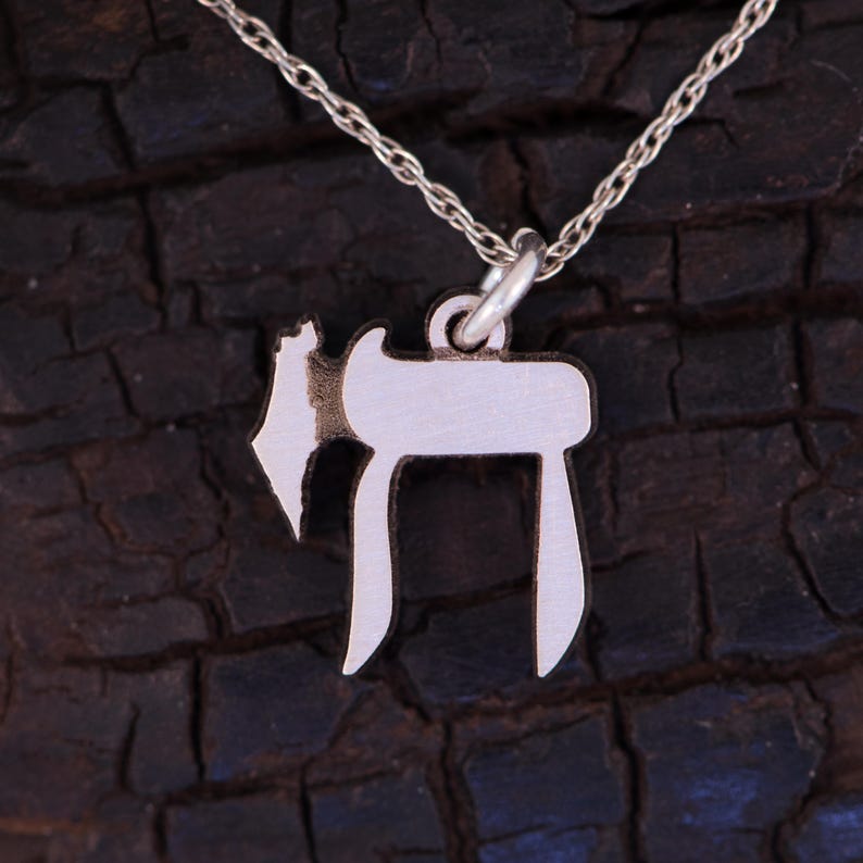 Puede incluir: Un collar de cadena de plata con un colgante de plata en forma de la letra hebrea Chai. El colgante est&aacute; sobre un fondo marr&oacute;n oscuro.