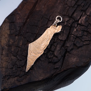 May include: A gold-toned metal pendant shaped like the map of Israel, featuring a textured, rugged surface finish. The pendant includes a small circular jump ring at the top for attachment, showcasing a detailed, uneven relief map design.