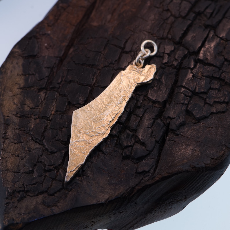 May include: A gold-toned metal pendant shaped like the map of Israel, featuring a textured, rugged surface finish. The pendant includes a small circular jump ring at the top for attachment, showcasing a detailed, uneven relief map design.