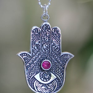 May include: A silver hamsa hand charm necklace with an intricate design and a pink gemstone in the center of the eye.