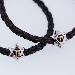 K&ouml;nnte beinhalten: Zwei schwarze Lederarmb&auml;nder mit silbernen Davidstern-Anh&auml;ngern. Jeder Anh&auml;nger hat einen goldenen hebr&auml;ischen Buchstaben in der Mitte.