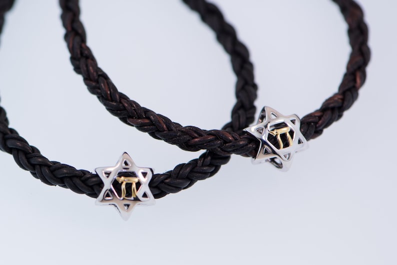 K&ouml;nnte beinhalten: Zwei schwarze Lederarmb&auml;nder mit silbernen Davidstern-Anh&auml;ngern. Jeder Anh&auml;nger hat einen goldenen hebr&auml;ischen Buchstaben in der Mitte.
