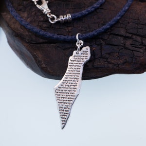 May include: A silver-colored pendant necklace featuring a detailed outline of a country, with text inscribed on the pendant. The pendant hangs from a dark blue braided cord necklace with a silver clasp. The necklace rests on a dark wooden surface.