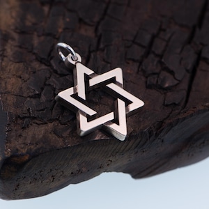 May include: A silver Star of David pendant charm with a detailed, intricate design. The charm is resting on a dark, textured surface.