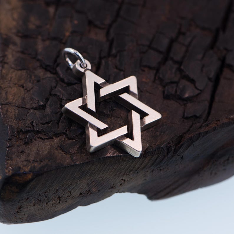 May include: A silver Star of David pendant charm with a detailed, intricate design. The charm is resting on a dark, textured surface.