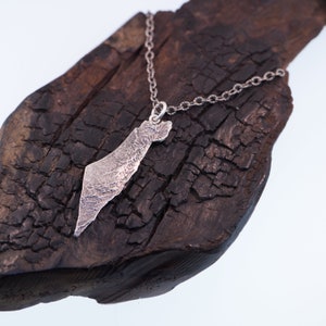 May include: A silver-toned metal pendant shaped like the map of Israel, featuring a highly textured, rugged surface. The pendant is suspended from a matching silver-toned metal chain link necklace, creating a distinct and detailed jewellery piece.