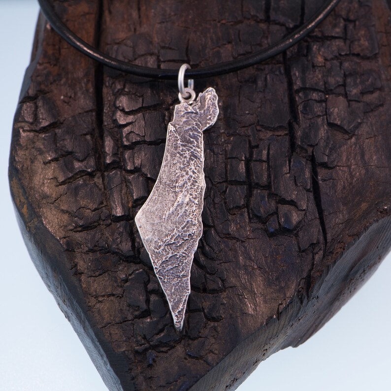 May include: A silver pendant shaped like the map of Israel with a textured, rugged surface finish. The pendant is attached to a simple silver jump ring and hangs from a smooth, thin black cord necklace, creating a contrast between the metal and cord.