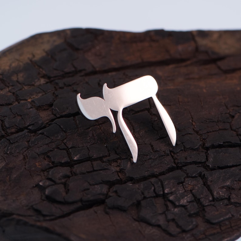 K&ouml;nnte beinhalten: Ein silbernes Chai-Symbol, das im Hebr&auml;ischen "Leben" bedeutet, liegt auf einer dunklen, strukturierten Holzoberfl&auml;che. Das Chai-Symbol ist eine stilisierte Darstellung der hebr&auml;ischen Buchstaben Chet und Yud.