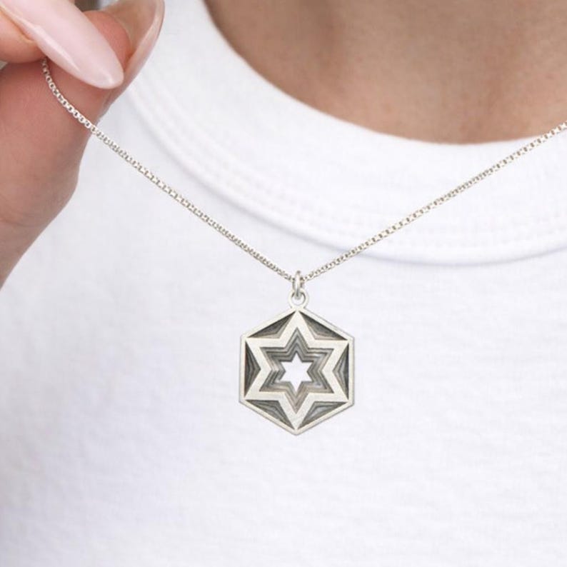 Peut inclure: Un collier argent&eacute; avec un pendentif hexagonal orn&eacute; d'un motif d'&eacute;toile superpos&eacute;e. Le pendentif pr&eacute;sente une &eacute;toile centrale entour&eacute;e de contours d'&eacute;toiles concentriques en nuances de gris. Le collier est pr&eacute;sent&eacute; sur fond blanc.