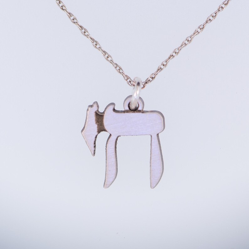 Puede incluir: Un collar de cadena de plata con un colgante de esmalte blanco en forma de la letra hebrea Jai.
