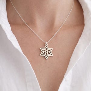 May include: A silver necklace featuring a Star of David pendant. The pendant has a six-pointed star design with an openwork pattern. The necklace is worn against a white shirt.