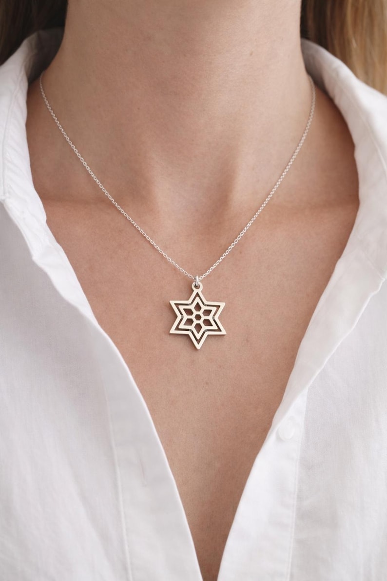 May include: A silver necklace featuring a Star of David pendant. The pendant has a six-pointed star design with an openwork pattern. The necklace is worn against a white shirt.