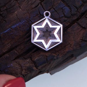 Peut inclure: Pendentif argent&eacute; avec un motif d'&eacute;toile superpos&eacute;e dans un cadre hexagonal. Le pendentif est suspendu &agrave; une petite boucle. Le fond est une surface en bois sombre et textur&eacute;e.