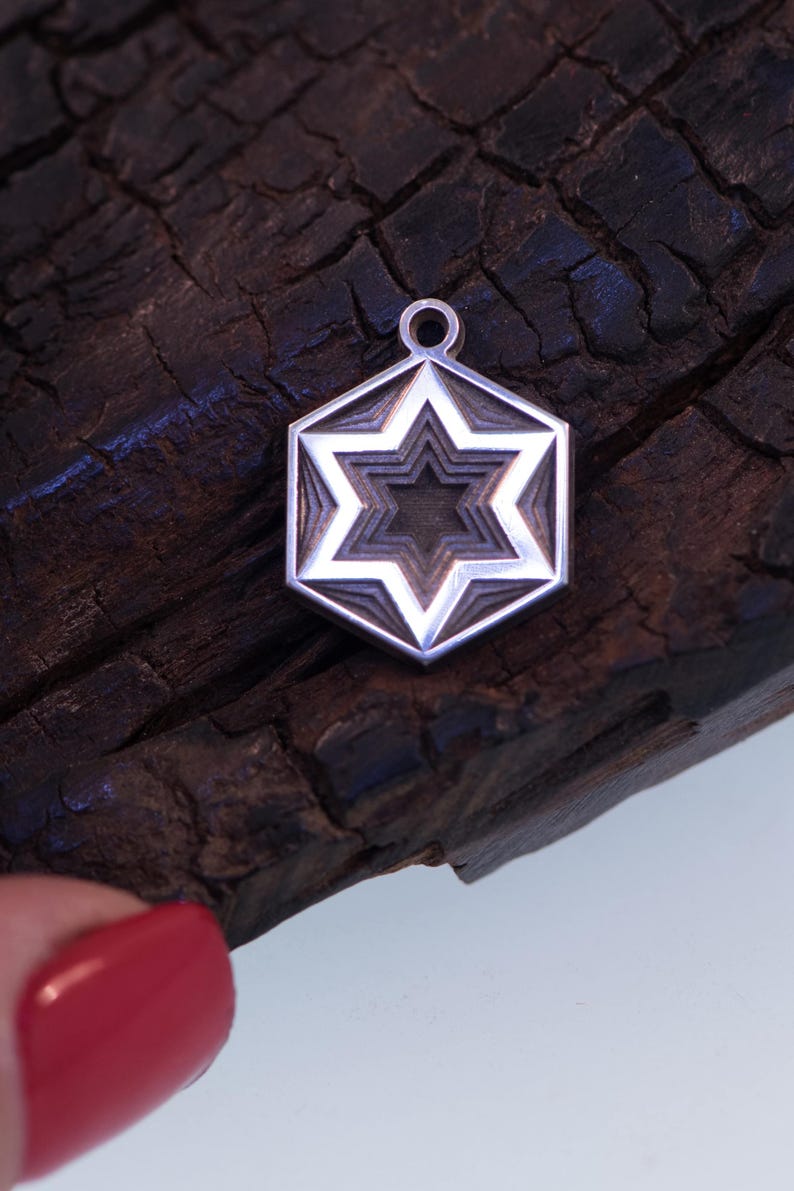 Peut inclure: Pendentif argent&eacute; avec un motif d'&eacute;toile superpos&eacute;e dans un cadre hexagonal. Le pendentif est suspendu &agrave; une petite boucle. Le fond est une surface en bois sombre et textur&eacute;e.