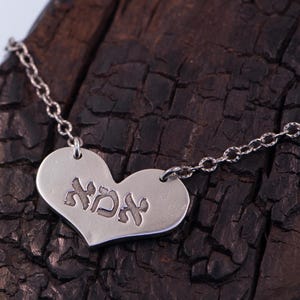 May include: A silver heart-shaped pendant necklace with the Hebrew word "אמא" engraved on the front. The pendant hangs from a delicate chain. The necklace is displayed on a dark, textured wooden surface.