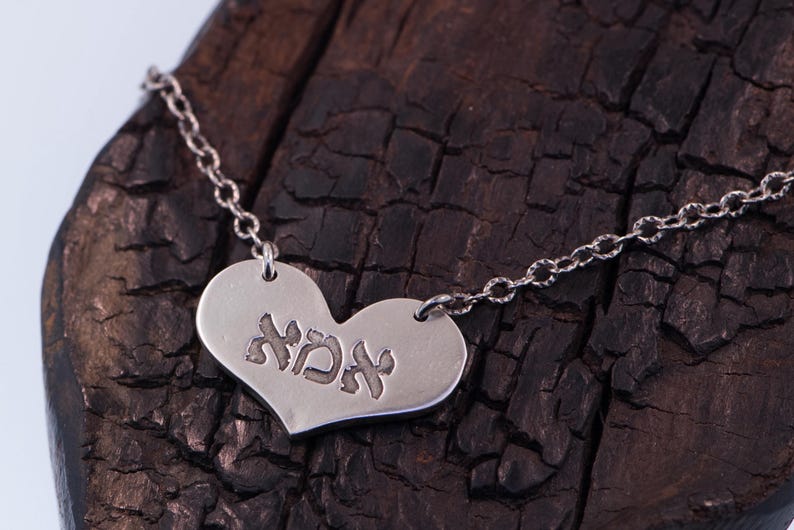 May include: A silver heart-shaped pendant necklace with the Hebrew word "אמא" engraved on the front. The pendant hangs from a delicate chain. The necklace is displayed on a dark, textured wooden surface.