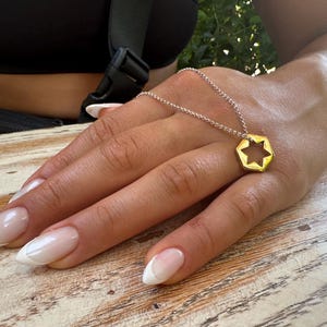 May include: A gold necklace with a hexagonal pendant featuring a star of David cutout. The necklace is worn by a person with white-tipped nails.